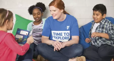 An image of a Caucasian female childcare provider at the YMCA reading with three young kids: an African American girl, a Caucasian girl, and a Hispanic boy. They are all sitting down on bean bags or on the colored-tile foam flooring.