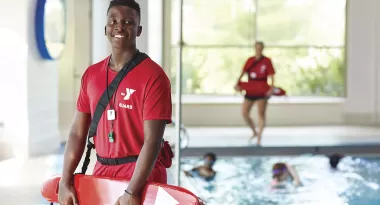 An image of an African American male lifeguard wearing a red YMCA branded t-shirt, and red buoy life-saving device. He is captured standing and smiling in front of the indoor YMCA pool. There are children with googles on and swimming in the pool that are visible in the image, too. There is also a female lifeguard wearing the same thing as the male, watching over the kids as they swim.