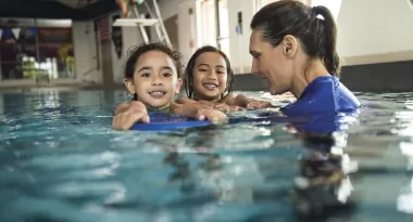 A Caucasian female YMCA swim instructor helps to teach two young girls how to kick as a swimming skill in the pool at the facility. 
