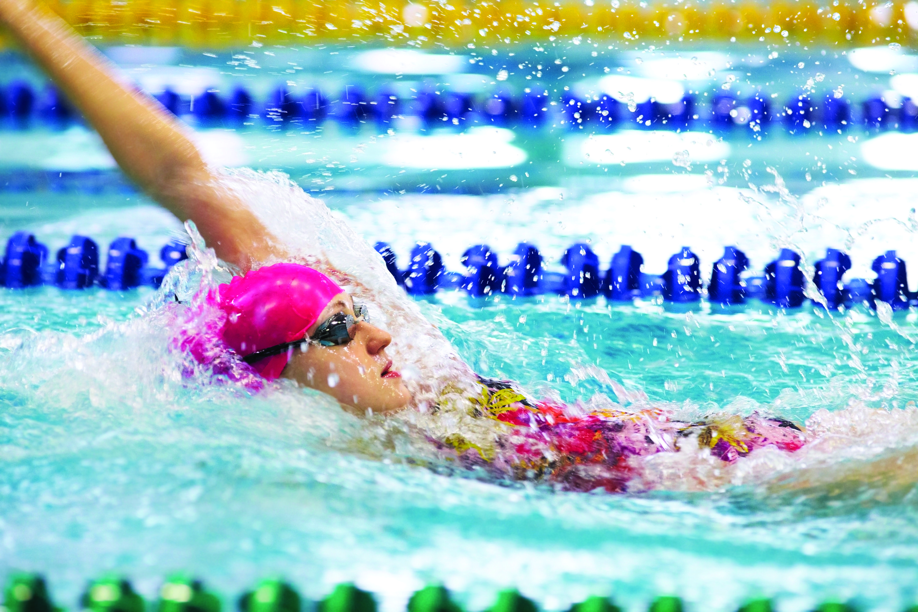 An image of a Caucasian female swimmer captured doing a backstroke swim in the lane of a pool at the YMCA. She is wearing a multi-colored swimsuit, pink cap on her head, and goggles. She has a determined look on her face.