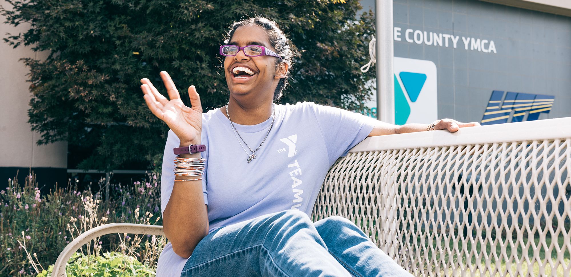 An image of an Indian teenage girl sitting on the bench outside of a St. Louis YMCA. She is wearing a purple t-shirt that says "Team Member," with the YMCA logo on it. She is waving at someone out of the frame, and smiling, widely.