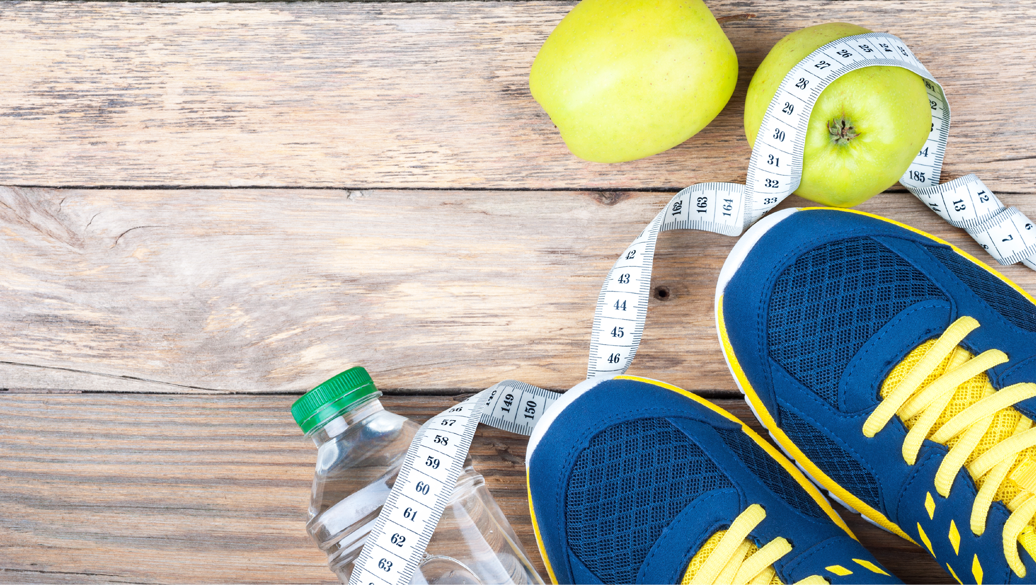 An image of a pair of navy blue and yellow tennis shoes standing on top of a twisted tape measurer and water bottle that are laying on a floor with wooden planks.