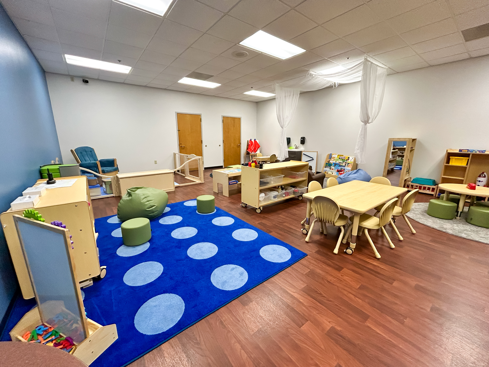 Wide view of the newly renovated YMCA Child Watch room featuring a blue accent wall, a colorful dotted rug, green bean bag chairs, and a gated infant play area.