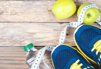 An image of a pair of navy blue and yellow tennis shoes standing on top of a twisted tape measurer and water bottle that are laying on a floor with wooden planks.