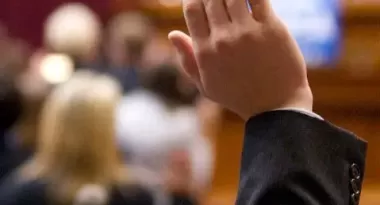 An image of a Caucasian male child's hand being raised during a meeting. He is captured wearing a black suit. The background is blurred, but it is visible that people are seated with their attention towards the speakers in the front of the room.