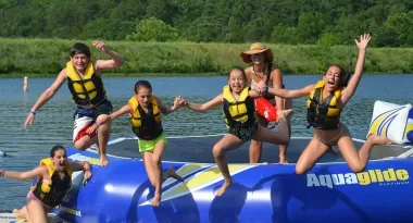 An image of a young group of Caucasian girls and one male jumping off of a water trampoline at Camp Lakewood. Three of the girls are holding hands whilst jumping in the water. Another female and the singular male are captured jumping in the left corner of the image. A lifeguard monitors them on the trampoline. All children are captured wearing life vests.