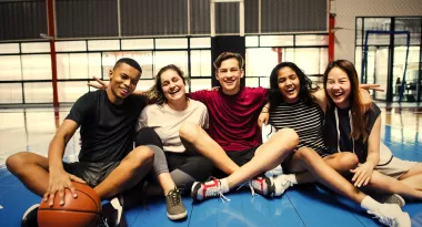 Five teenagers from the YMCA's teenage program pose for an image sitting arm-in-arm on a basketball court after a pick-up game. There are two teenage males captured, one African American male and one Caucasian male. There are also three teenage females captured, one Caucasian female, one Asian female and one African American female. All are smiling for the image.