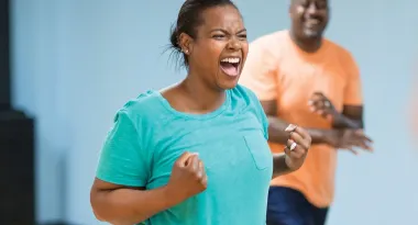 An image of an African American woman posing triumphantly with her fists closed, like she is excited to workout. An African American male is captured smiling at her in the background. On the right side of the frame, a Caucasian female is barely visible.