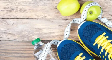 An image of a pair of navy blue and yellow tennis shoes standing on top of a twisted tape measurer and water bottle that are laying on a floor with wooden planks.