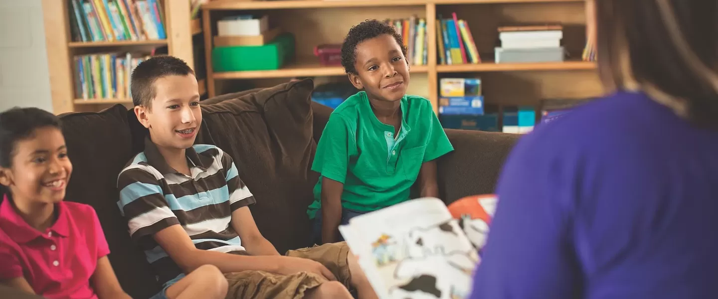 A female YMCA staff member wearing a purple shirt reads a book to three children, one Hispanic girl, one Caucasian boy, and an African American boy. All three children are happily listening to the staff member read to them while they sit on a couch in their classroom.