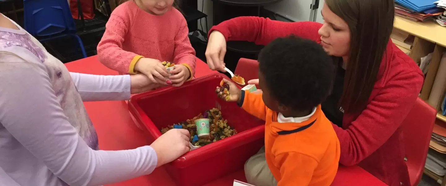 An image of a Caucasian female staff member at the YMCA helping three after school children, two Caucasian girls and one African American boy, ranging in ages. They are all playing with a box filled with small figurines.