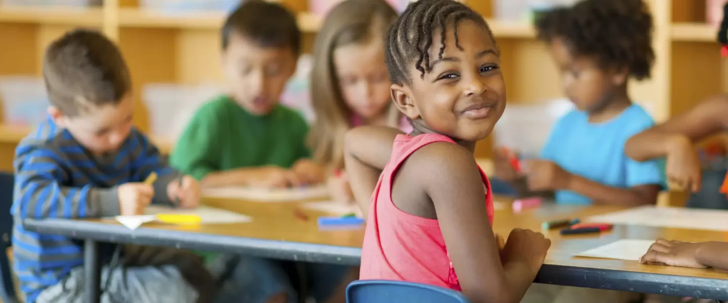 A young African American girl poses looking back towards the camera with a soft smile on her face. She is captured sitting, coloring pieces of papers with colorful markers with a diverse group of her classmates at a table in a classroom at the YMCA.
