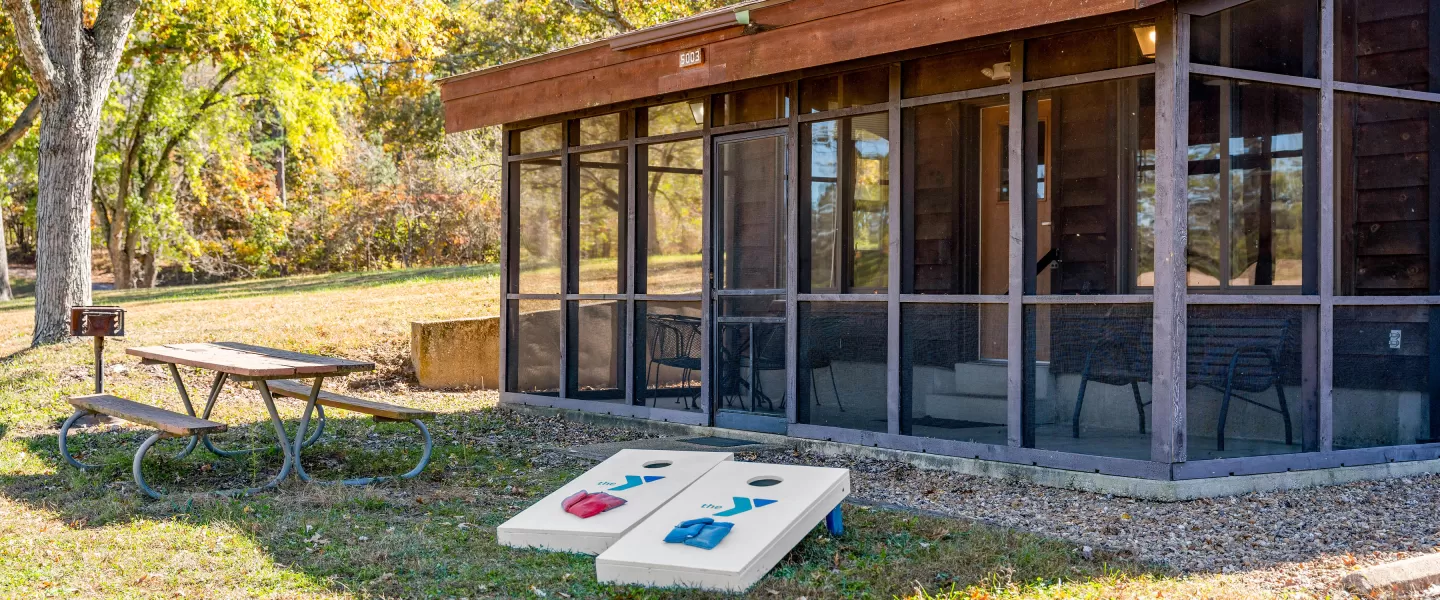 Bluff View Cabin exterior with screened-in porch, picnic table, charcoal grill, and YMCA-branded corn-hole game boards.