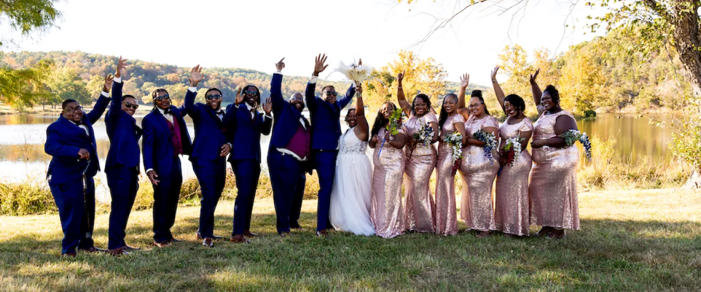 The full wedding party posing for a happy group photo by the lake at a sunny Trout Lodge wedding.