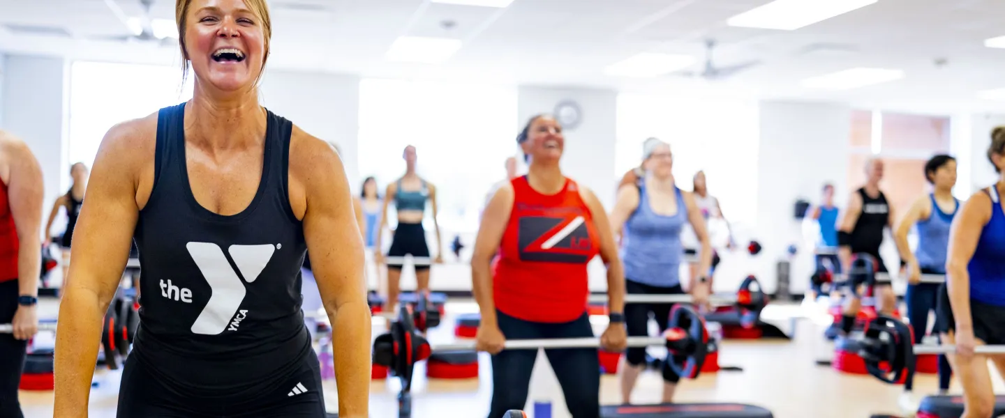 Les Mills Bodypump YMCA Group Exercise Class participants smile next to BODYPUMP steps holding the class weight bars