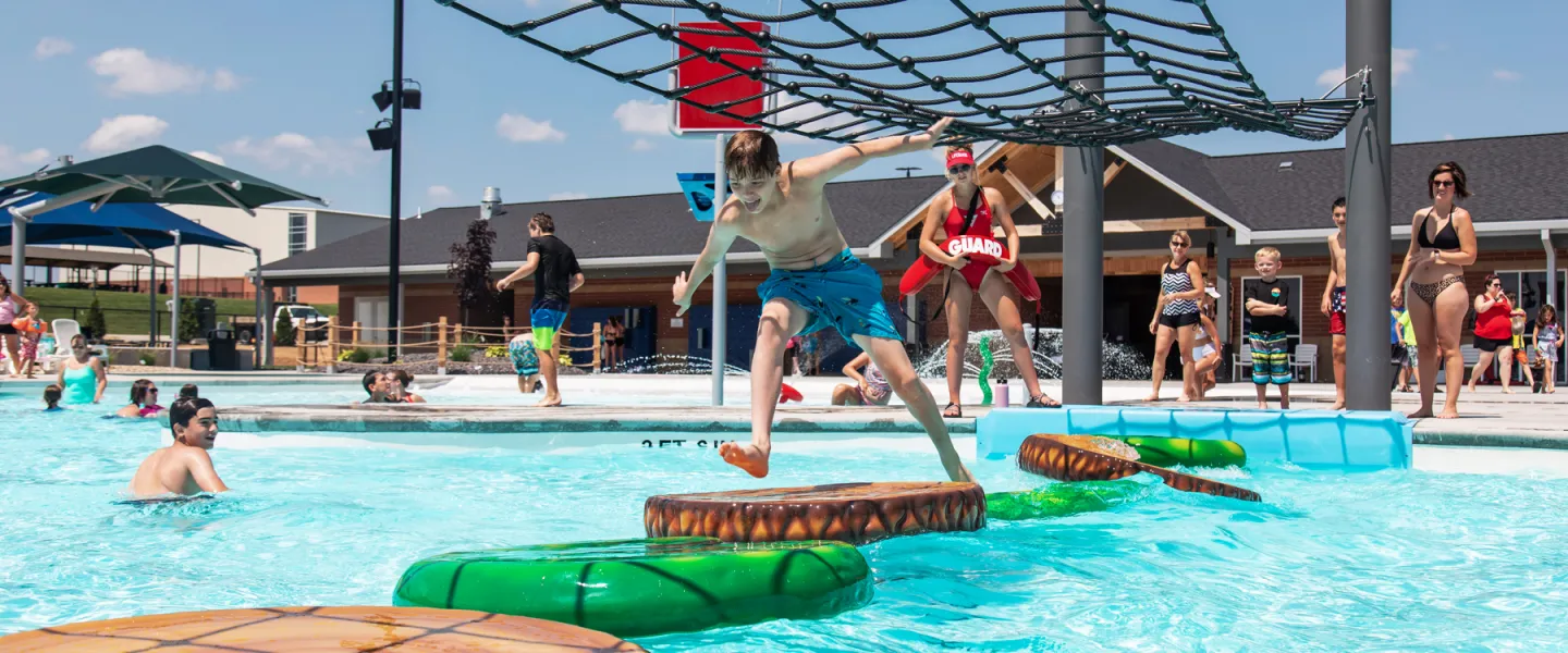 A young swimmer balancing carefully across the floating lily pad water obstacle course using the overhead rope net at the River Rapids Waterpark.