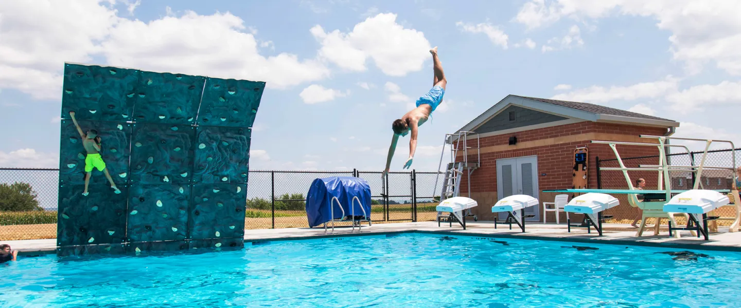 An adventurous swimmer diving backward into the deep end of the pool after climbing the poolside aquatic rockwall feature.