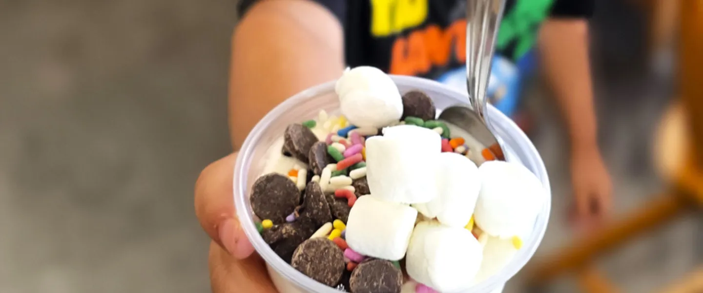 A young child holds a cup of ice cream with marshmallows and cookies to the camera with an excited look on his face