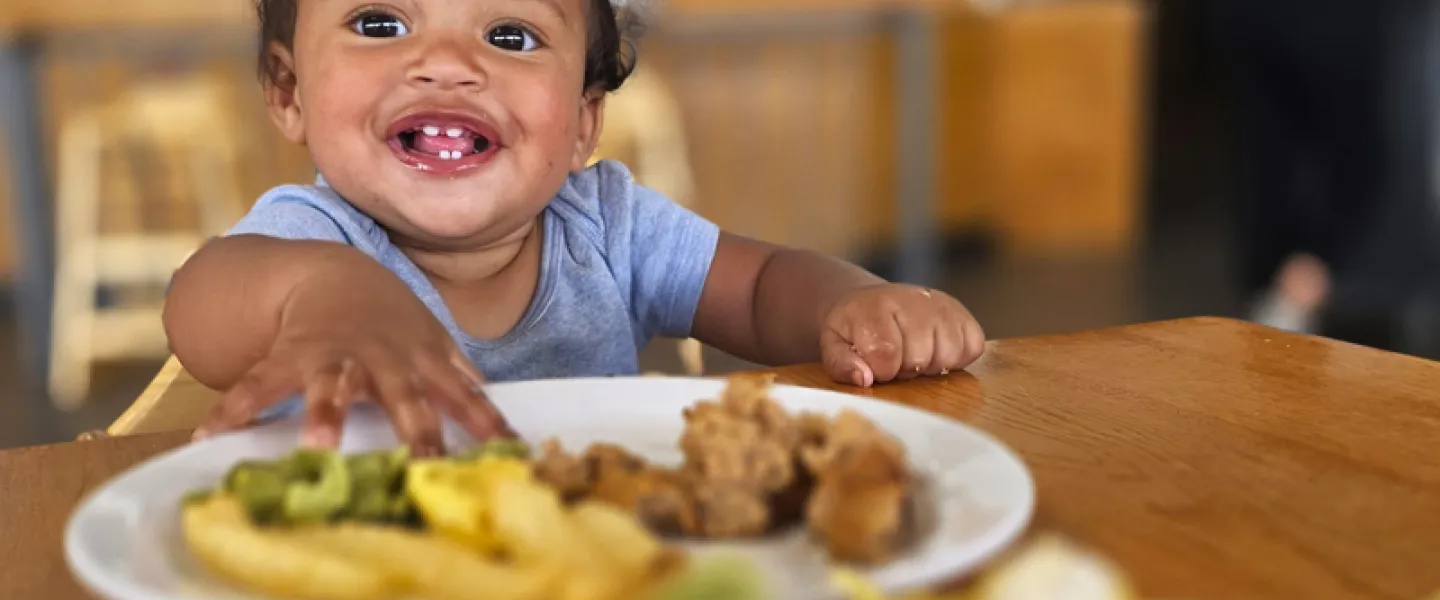 A smiling baby grabs for food on their plate while eating in the Dining Hall at YMCA Trout Lodge
