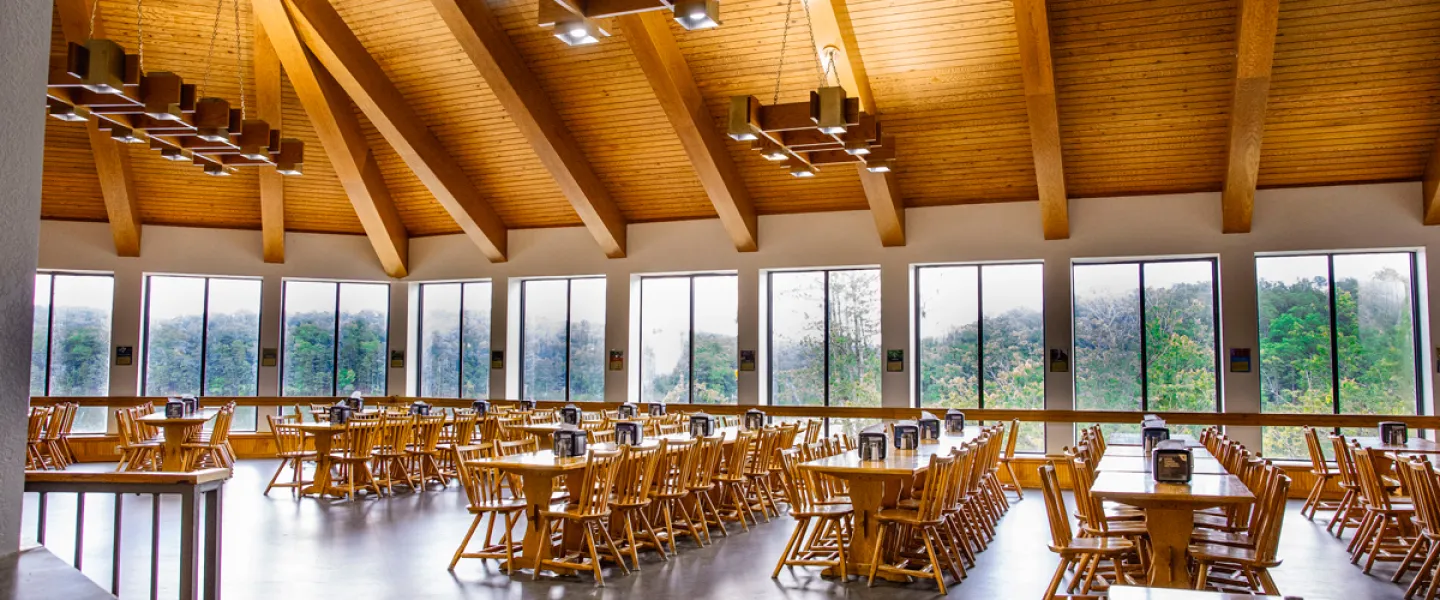 Interior view of the Trout Lodge Dining Hall showing rows of dining room tables and chairs next to wrap-around windows showing Sunnen Lake and Treetop views