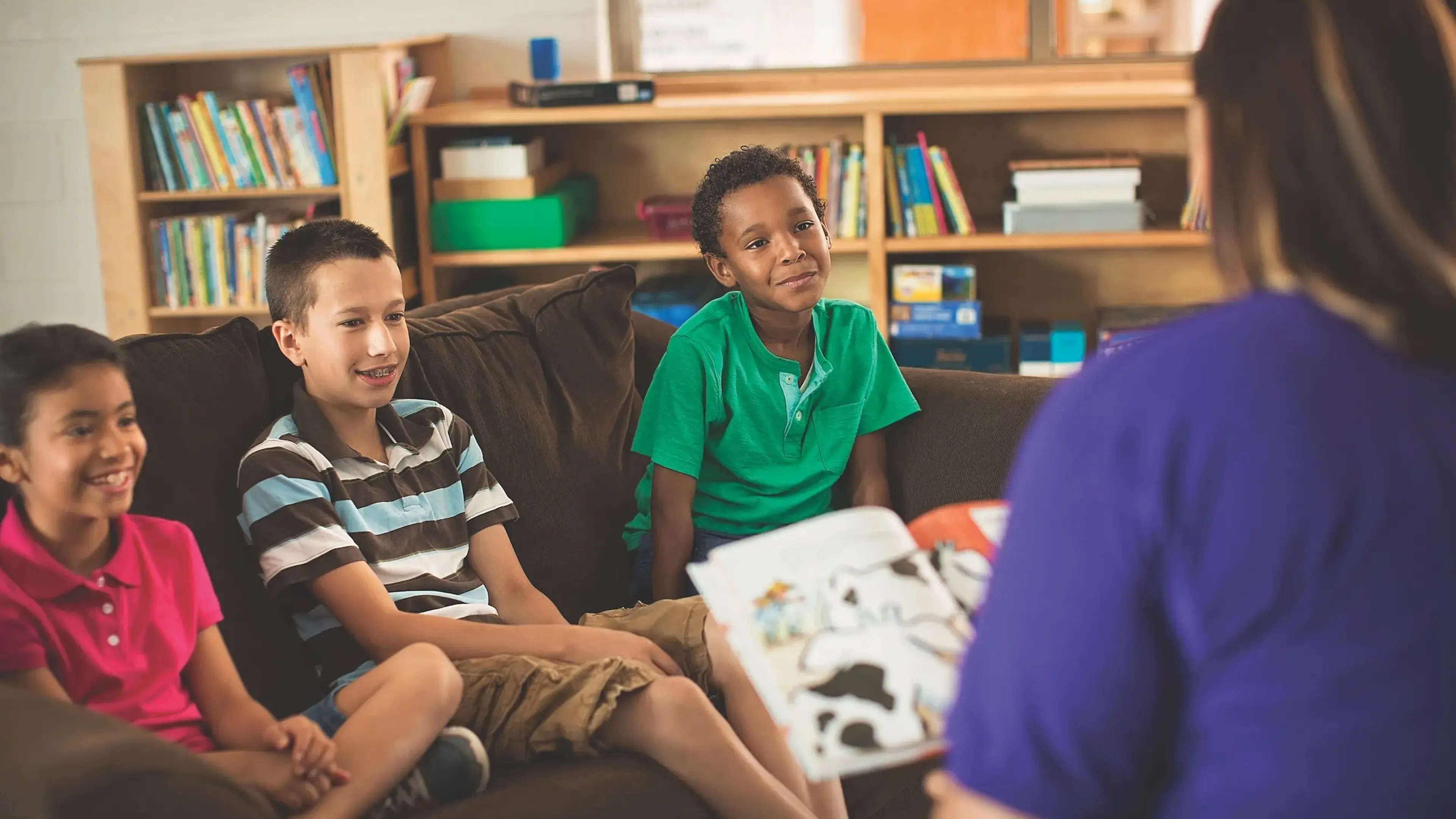 A female YMCA staff member wearing a purple shirt reads a book to three children, one Hispanic girl, one Caucasian boy, and an African American boy. All three children are happily listening to the staff member read to them while they sit on a couch in their classroom.