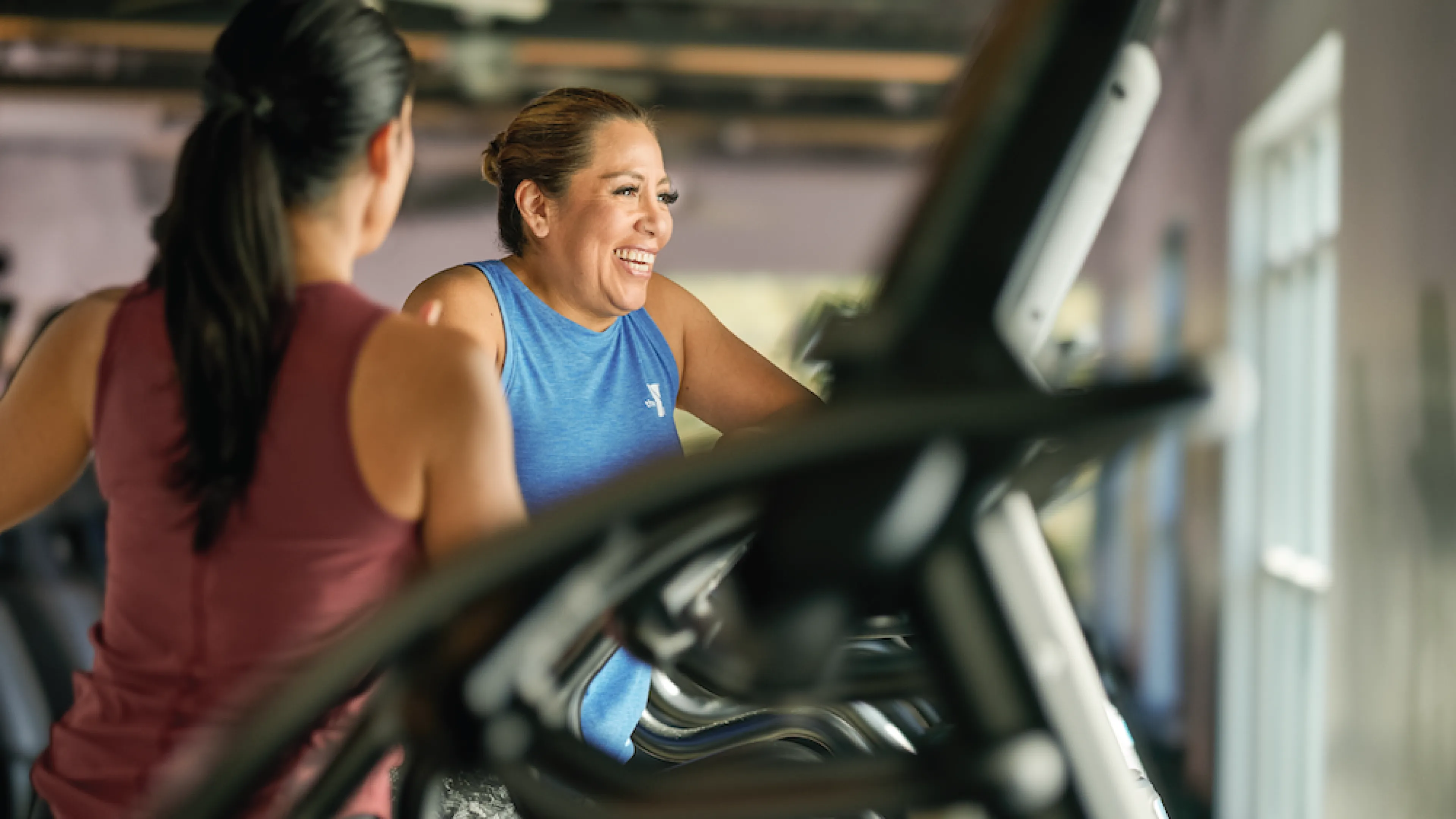 Two females talking and smiling as they work out on elliptical cardio machines in a YMCA Fitness Center.