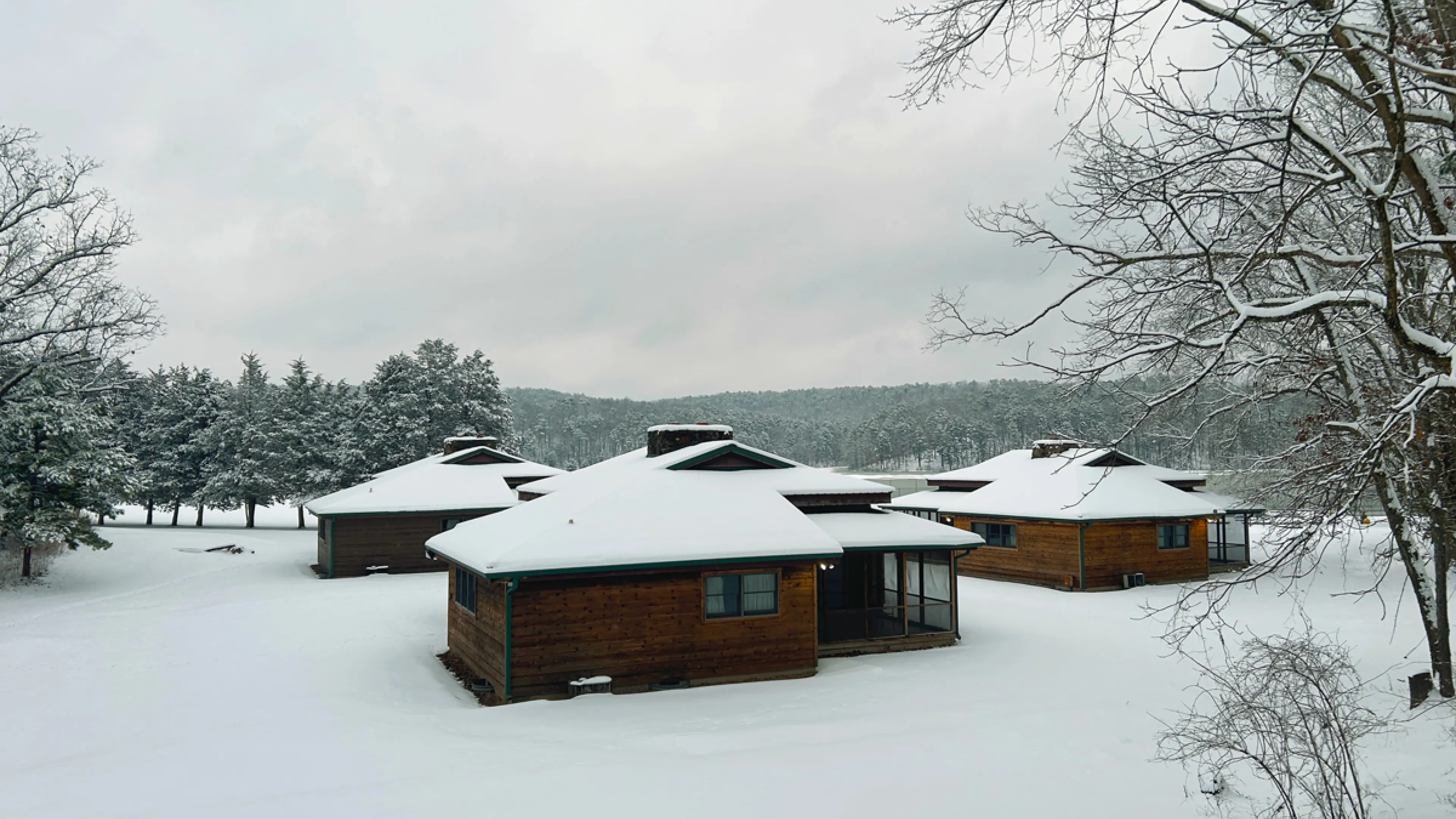 YMCA Trout Lodge Lake View Family Cabins covered in white winter snow next to the Ozark Hills evergreen trees covered in snow and a frozen Sunnen Lake