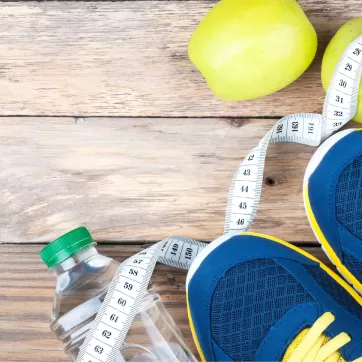 An image of a pair of navy blue and yellow tennis shoes standing on top of a twisted tape measurer and water bottle that are laying on a floor with wooden planks.