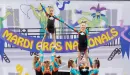 Thumbnail: Nine Caucasian cheerleaders from a YMCA team pose in a group stunt including two pyramids at their local cheer competition. The girls are dressed in the same black, blue, and orange uniforms.