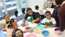 Thumbnail: An image of a Caucasian woman wearing an apron assisting a group of young African American boys and girls during an after school program provided by the YMCA. They are cooking and learning about the importance of nutrition.