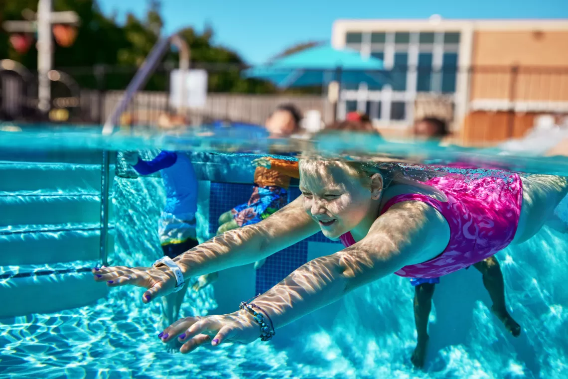 Girl swimming underwater through clear blue water in a YMCA outdoor swimming pool.