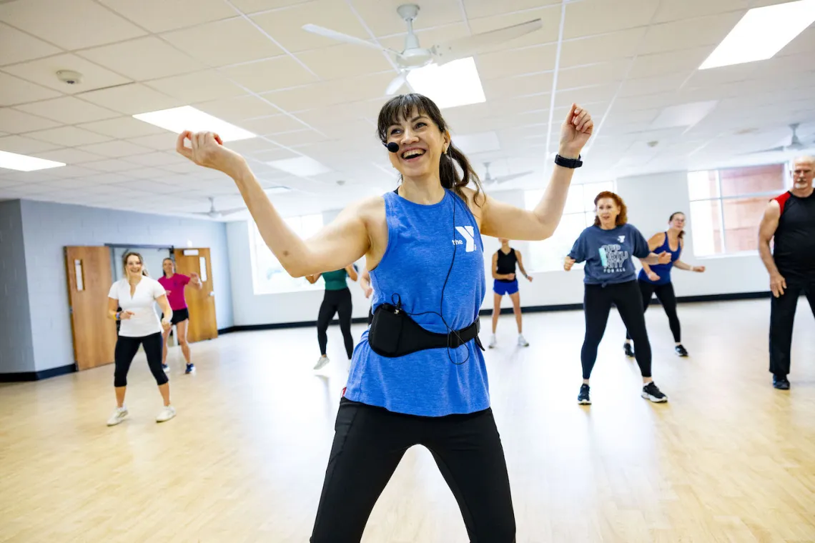 A happy YMCA Zumba Group Fitness Instructor leads a Zumba class in a bright group exercise studio with participants in view behind her.