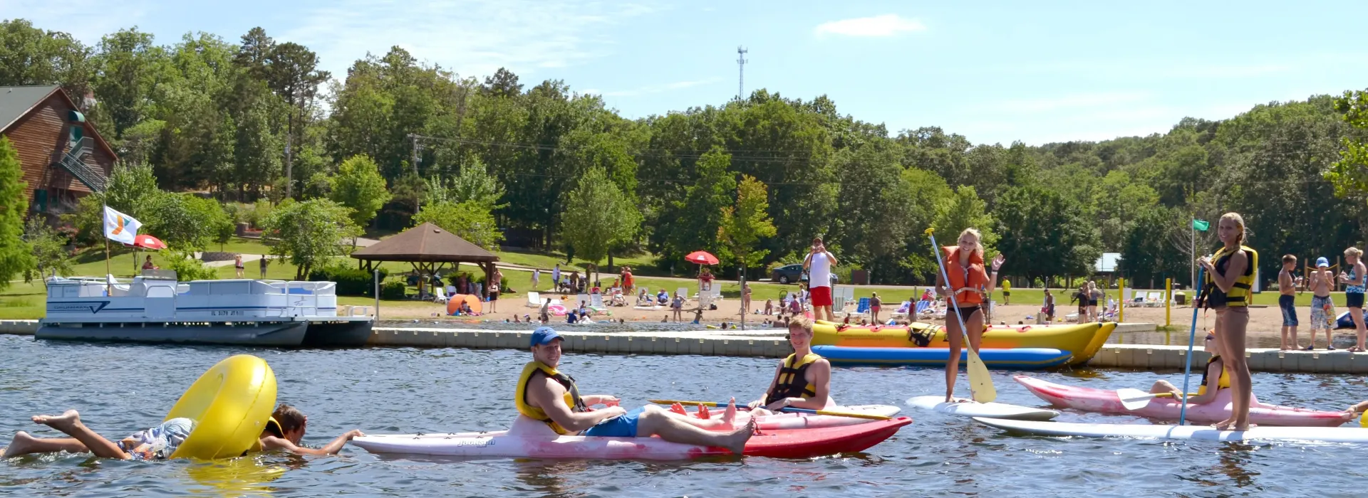 A group of male and female teens enjoy the lake and water-based activities during a program at Trout Lodge YMCA. The students are doing a variety of activities like kayaking, paddle-boarding, and swimming in inflatable floaties. All teenagers look to be having a fun experience.