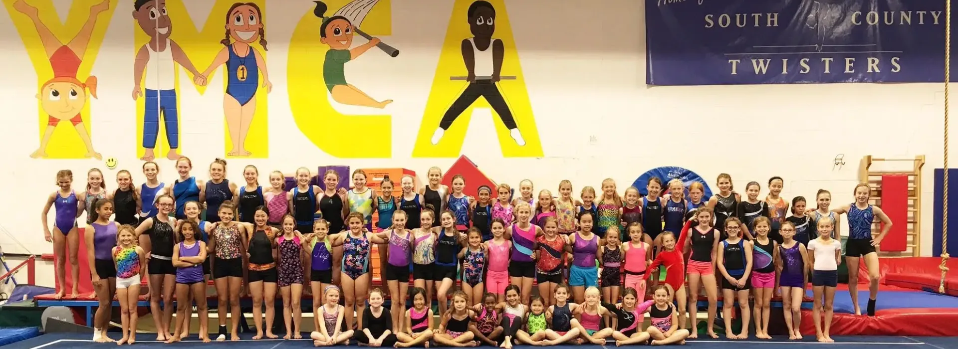 An image of the entire South County YMCA Gymnastics team. All ages and levels are present. The image was taken in the gymnastics and tumbling room in front of the wall with the word YMCA painted onto the wall.
