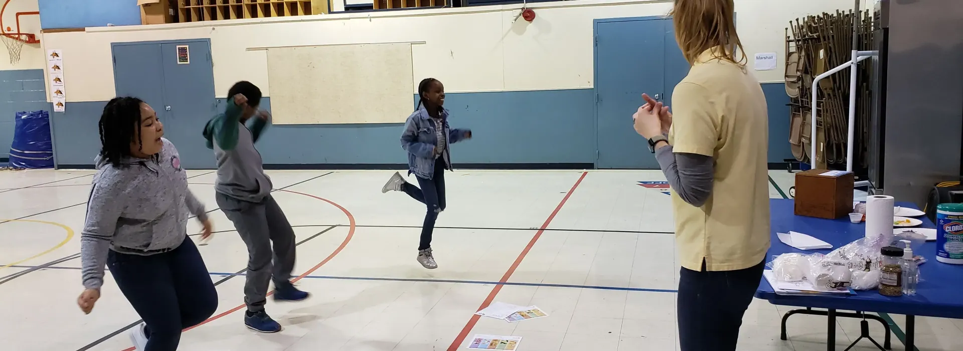 An image of a Caucasian after school leader playing a game where the three African American children in her care are captured jumping on one foot, smiling.
