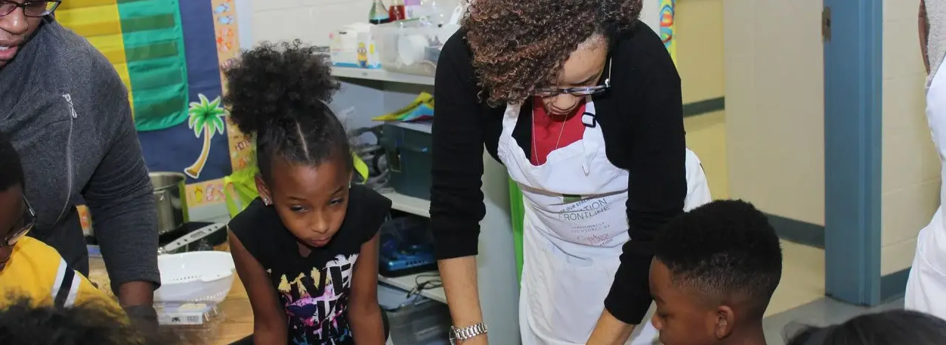 Two African American females in their middle ages helping a classroom of kids with nutrition skills. The instructor on the right is wearing a white apron over her clothes.