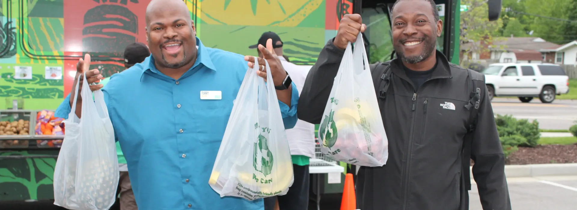 An image of two African American men posing in front of the YMCA's Metro Market Bus. They are enthusiastically holding up bags of food in their hands.