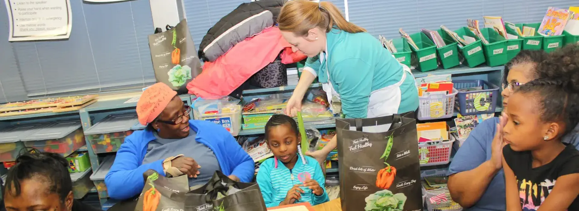 An image of a Caucasian woman from the YMCA helping to teach African American mothers and daughters essential nutrition and cooking skills in a classroom.