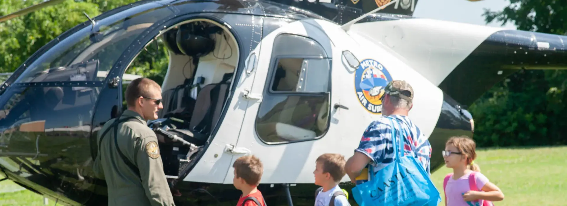 An image of three Caucasian kids standing in front of a police helicopter with the pilot and their father, during the backpack give-away at the YMCA.
