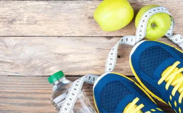 An image of a pair of navy blue and yellow tennis shoes standing on top of a twisted tape measurer and water bottle that are laying on a floor with wooden planks.
